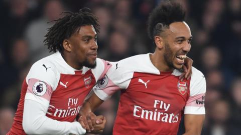 Alex Iwobi puts his left arm around the shoulder of Pierre Emerick-Aubameyang as both players celebrate a goal for Arsenal. Both players are wearing red and white Arsenal home shirts