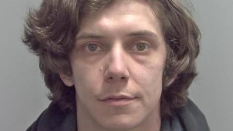 A police custody image of a man staring blankly at the camera. He has thick wavy brown hair and hazel eyes.