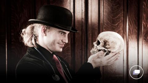 A picture of a male with long blonde hair tied up, in a black top hat holding a skull. 