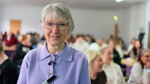 Joan Lyon, a 75-year-old woman, with grey bobbed hair and glasses, wearing a lilac cardigan. Behind her are groups of students sitting at tables..