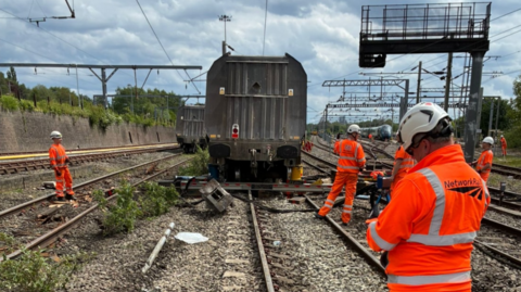 A number of men in orange reflective clothing and white helmets standing on railway lines with a large carriage in front of them