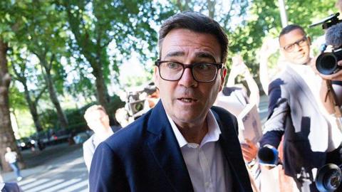 Andy Burnham flanked by reporters and photographers on Millbank. He is wearing a white shirt with a black jacket and glasses. There are trees in the background.