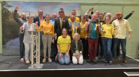 The Lib Dems celebrate in Witney - various members on a stage, some wearing yellow T-shirts or ties. All ae smiling and some have their hand and arms in the air in celebration
