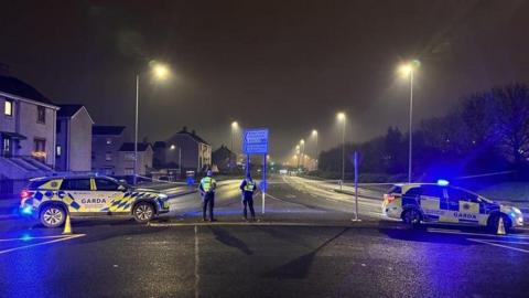 Two Garda police cars block off the entrance and exit to a road. Two officers look down the empty road. IT appears to be at the exit to a roundabout. There is a housing development to one side of the road, and trees to the other. 