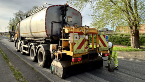 A highway maintenance vehicle on a road with a man wearing a white hard hat and a fluorescent outfit holding a pipe into a bucket