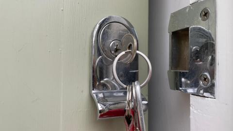 A silver key on a keyring sits in the lock on a white door. The latch has been opened. 