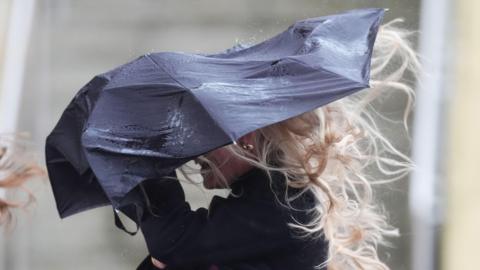 A woman with long blonde hair takes shelter under a navy blue umbrella during stormy conditions. Her hair is blowing behind her. He umbrella has been bent out of shape.