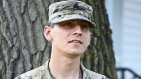 Soldier in a military cap and fatigues stands in front of a tree.