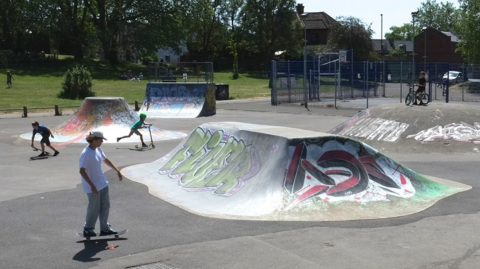 A skate park with two boys using skateboards and another on a scooter. There are skate ramps which have graffiti tags on them. In the background people are sat on some grass under trees.