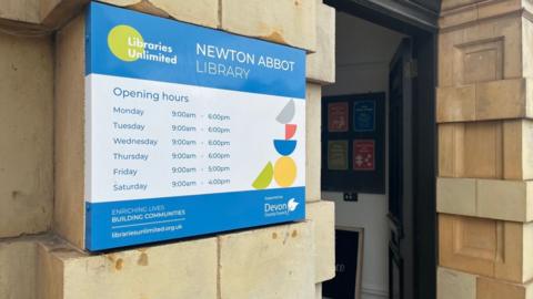 The opening hours sign at Newton Abbot libraryin blue and white on a Portland stone wall