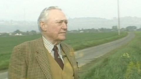 Richard Todd stand in a tweed jacket, mustard wastecoat maroon tie and white checked shirt, look contemplatively out over a field in Normandy.