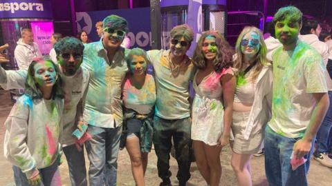 Eight young adults stand in an outdoor space with their arms around each other. Their shirts are covered in coloured powder. 