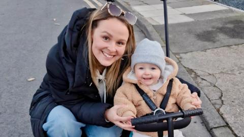 Alice Cole crouches down by her baby boy Oliver, both of whom are smiling at the camera. Alice is wearing a coat and jeans with sunglasses on her head, while Oliver is in a hooded fleece with a grey hat on his head.