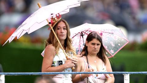 Two people stand looking in the direction of the camera while holding pink umbrellas with a bokeh background, while at the York races on 20 August.