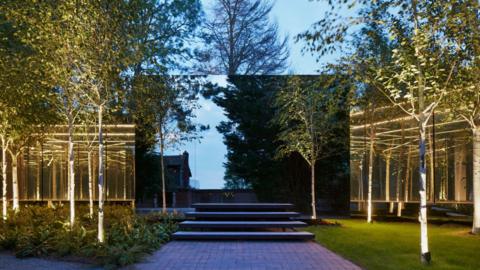 A building that looks like a large black television screen with steps leading up to it. Trees and yellow lights surround the house. 