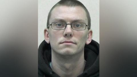 Mugshot of James Gregory. He is clean shaven with short dark hair and is wearing glasses and a black hoody.