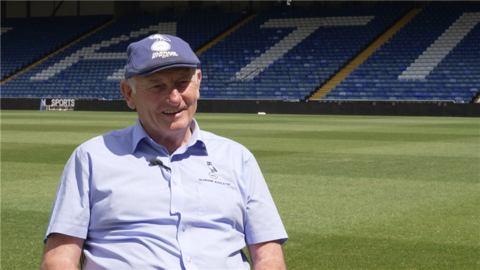 Frank Rothwell is wearing a light blue shirt and a navy flat cap. He is sat next to the Oldham Athletic pitch. 