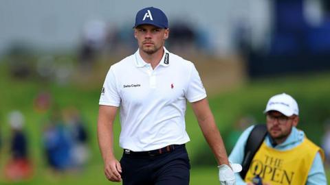 Curtis Knipes walking on the Portrush course, wearing a white polo shirt, dark trousers and a dark blue baseball cap with an 'A' logo. His caddie is pictured in the background