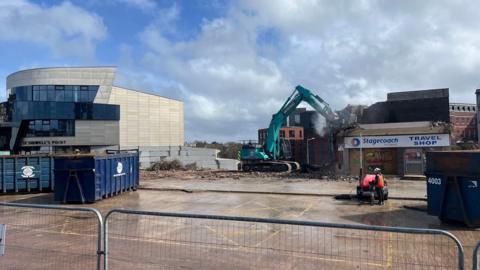 A digger demolishing a building on the site of the old coach station in Exeter with St Sidwell's Point leisure centre in the background
