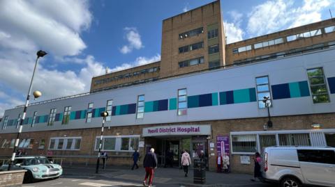 The picture shows the entrance to Yeovil District Hospital.  There are people walking on a crossing.  There are two vehicles outside the hospital.