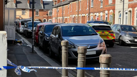 A general view picture of a street off London Road in Reading, with cars packed alongside the road and a police car parked in the middle of the road. There is police tape cordoning off the street close to the camera.