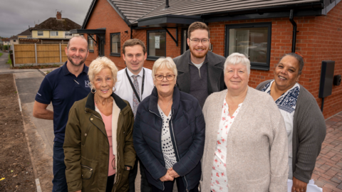 Outside a new bungalows in School Lane are back row - Nathan Bayliss, Keon Homes Site Agent, George Williams, Wolverhampton Homes Project Manager, Councillor Chris Burden, Cabinet Member for City Development, Jobs and Skills, Teresa Phillips, resident and Wolverhampton Homes Board Member.  Front - Rose Davis, resident, Diane Brookshaw, Tenants and  Residents’ Association Chair, and Jackie Swift, Resident and Tenants and  Residents’ Association Committee Member.
The men and women are stood on a path right outside a new red-bricked home with some land yet to be landscaped on the left of the picture.