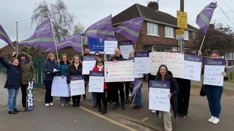 The photo shows a group of people smiling for the photo. They're each holding vairous signs and purple flags which read UNISON on them. 