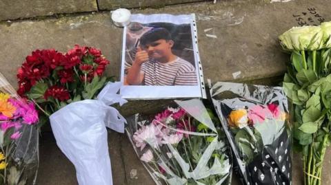 A number of floral tributes to the four Shrewsbury teenagers who died in the crash in November 2023. There is also a picture of one of the victims with his thumb up, when he was younger, in a plastic envelope.