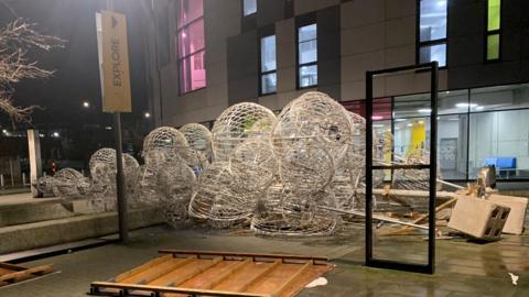 The silver Christmas tree which is made up of several large baubles is pictured lying on its side on the ground after it fell over in strong winds. Concrete blocks that had been holding it up have also fallen over. Other debris can be seen scattered around the tree in the night time image. 