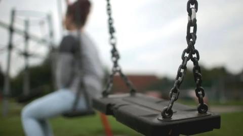 A blurred out picture of a girl sitting on a swing in a playground, with her back to the camera, wearing faded blue jeans and her hair in a bun.