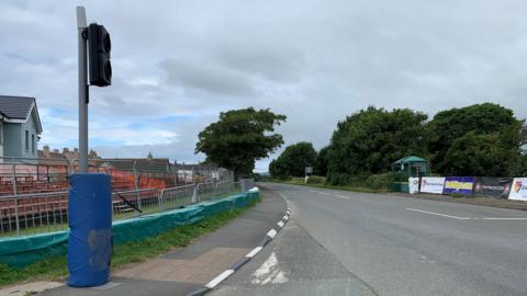 The road at Castletown Corner on the Southern 100 course. There are banners on the right and a small orange grandstand on the left behind fencing and barriers. There is a blue padding around the bottom of a traffic light pole.