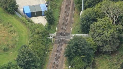 An aerial view of a railway line cutting through green woodland, with a fenced pedestrian crossing over the tracks and several coloured storage containers on a paved area nearby.