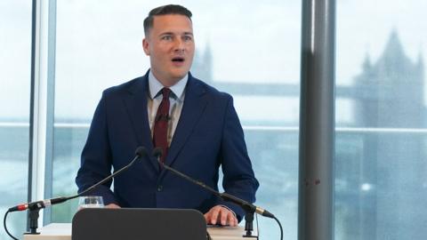 Wes Streeting wearing a navy suit and a red tie. He is standing at a lectern on front of a view of Tower Bridge in central London