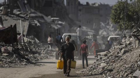 A Palestinian man carries water cans among the rubble of destroyed buildings on the streets of Sheikh Radwan, Gaza City (20 October 2025)