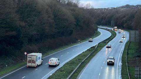 The A38 dual carriageway in Plymouth. There are cars on both sides of the road with headlights on. 