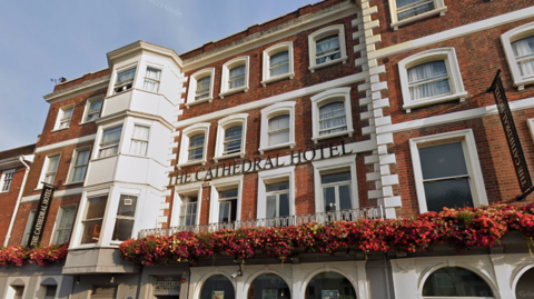 A google maps street view of The Cathedral Hotel in Salisbury. The building is red brick with white details. There is a row of hanging flowers across the width of the building. There are many windows and a sign reading "The Cathedral Hotel" in capital letters. 