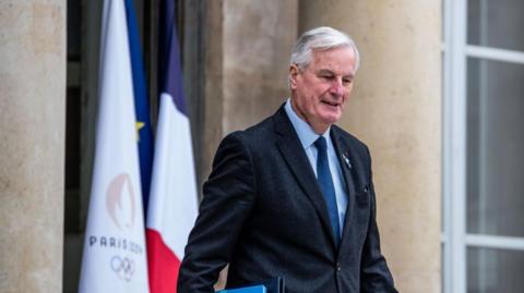 Michel Barnier walks in front of a French flag. He is wearing a black jacket and a blue tie.