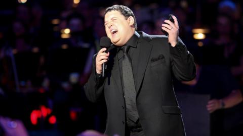 Peter Kay wearing a black suit preforming a gig, holding a microphone and laughing 