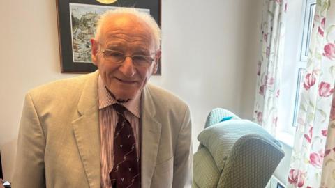 An older man with short grey hair, wearing glasses and smiling at the camera. He is wearing a beige suit jacket.