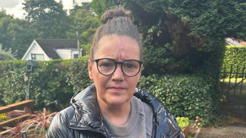 Charmaine standing outside wearing a grey jumper and black coat. Her black hair is tied up and she is wearing black framed glasses. 