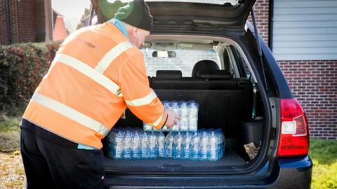 A man in an orange hi-vis jumper taking out slabs of bottled water out of a black vehicle. 