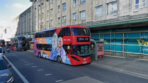 A red double decker bus parked up by a bus shelter outside the Poundland store on Royal Parade in Plymouth. Scaffolding has been erected outside the shop. The bus has a large advert for St Luke's Hospice on its side which features a male and female hospice worker.