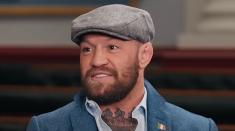 McGregor is wearing a grey flat cap, a blue suit, white shirt. There are tattoos on his neck.He is smiling.