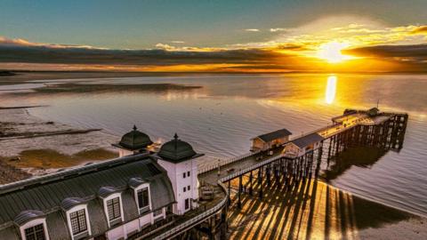 The bright yellow sun sets over water with a building and pier in the forefront