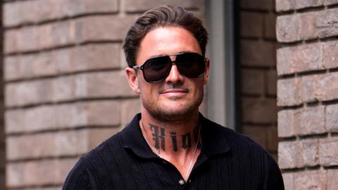 Stephen Bear smiles at the camera as he walks in to court, with a brick wall behind him. He has dark hair and stubble and is wearing big sunglasses and a black shirt. He has a prominent tattoo on his neck.