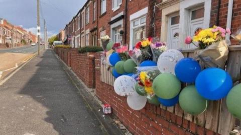 Balloons and bunches of flowers tied to a house's front wall at Park Road, Stanley. 