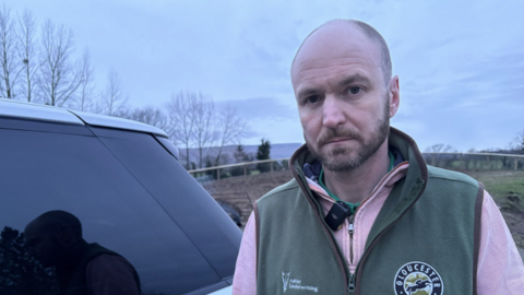 A bald man with a short beard stands next to a silver 4x4 wearing a pink quarter zip fleece and a green gilet.