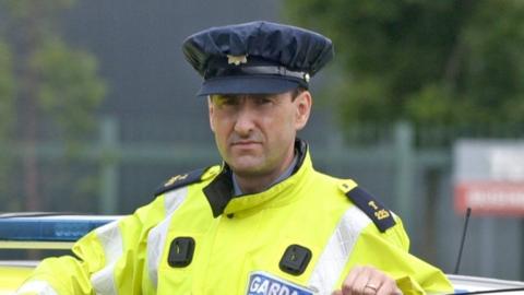 A man wearing an Irish police officer uniform of a yellow hi-vis jacket standing behind the open door of a police car which is stationary.
