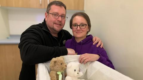 A man in a black Forever Stars sweatshirt put his arm around his wife who is wearing a purple Forever Stars hoodie. They are crouched down next to an empty cot with two teddy bears inside.