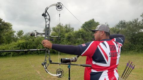 An archer wearing a Team GB with GBR Scott written on the back over a Union Jack kit draws back his bow preparing for a shot at target.